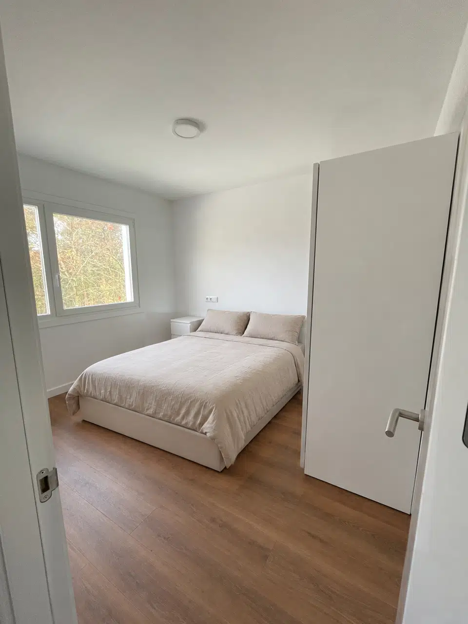 Dormitorio principal con cama doble, mesilla blanca y ventana al arbolado