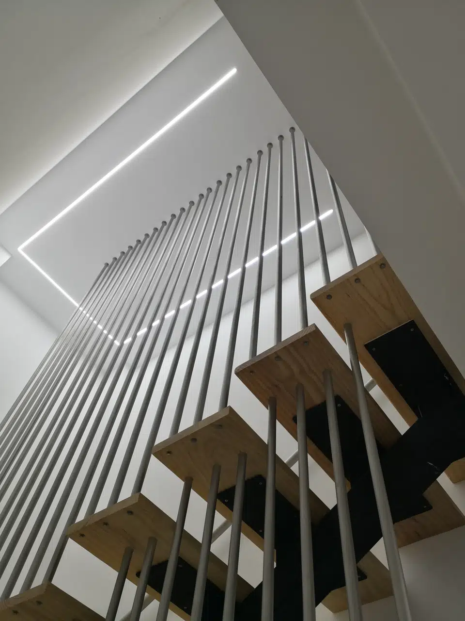 Vista ascendente de la escalera con peldaños de madera, varillas metálicas e iluminación lineal